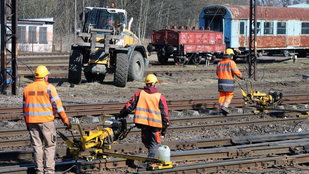 Pracownicy w odblaskowych kamizelkach i kaskach pracują na torach kolejowych podczas modernizacji infrastruktury.