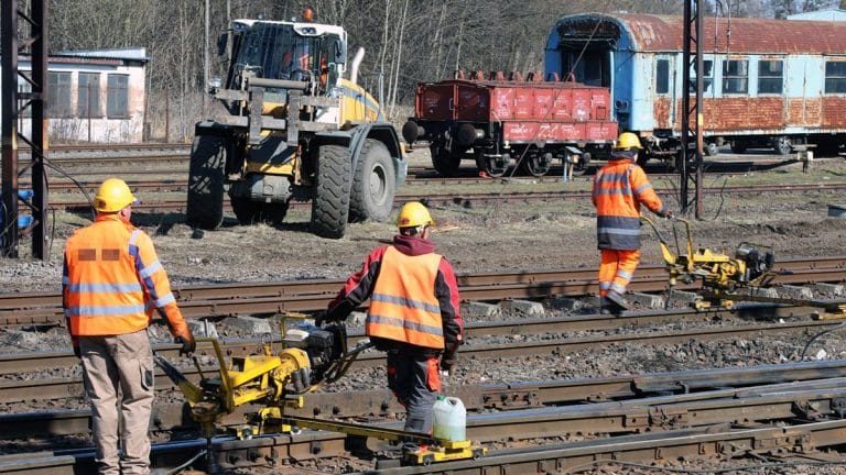 Pracownicy w odblaskowych kamizelkach i kaskach pracują na torach kolejowych podczas modernizacji infrastruktury.