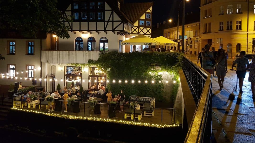Wieczorny widok na ulicę z restauracją i spacerującymi ludźmi, oświetlony lampkami i latarniami, w tle budynki.