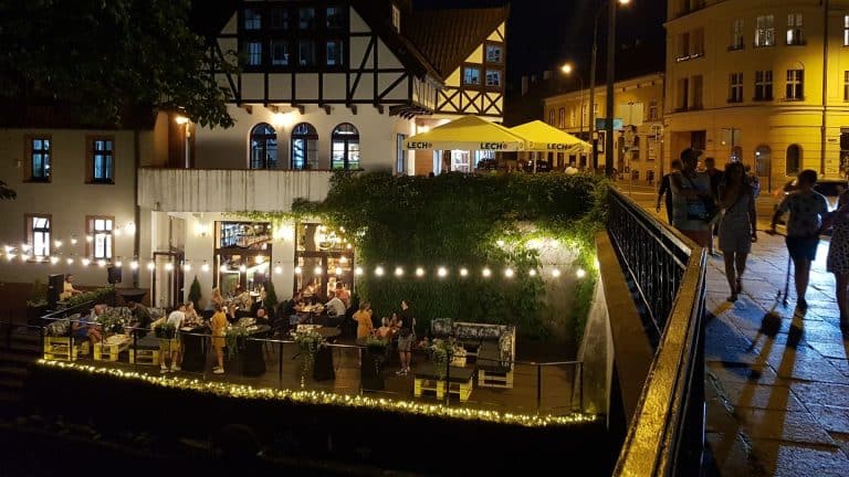 Wieczorny widok na ulicę z restauracją i spacerującymi ludźmi, oświetlony lampkami i latarniami, w tle budynki.
