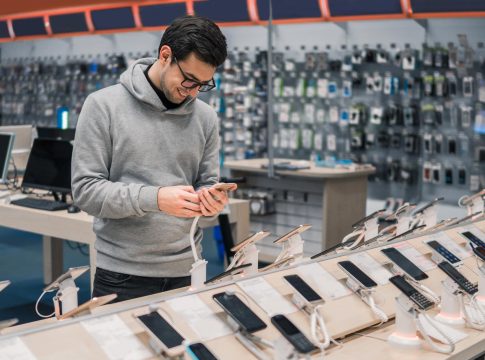 Mężczyzna w sklepie z elektroniką, przegląda telefon na tle wyświetlaczy i urządzeń na półkach.