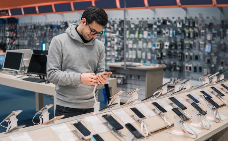 Mężczyzna w sklepie z elektroniką, przegląda telefon na tle wyświetlaczy i urządzeń na półkach.