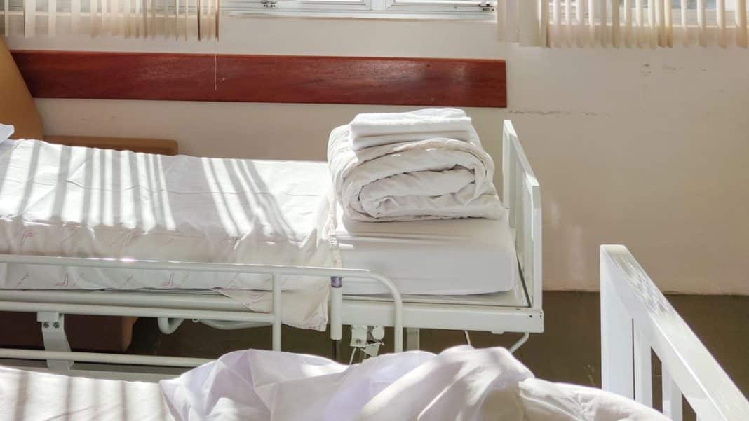 Pusty szpitalny łóżko z złożonymi białymi pościelami, w tle okno z żaluzjami.