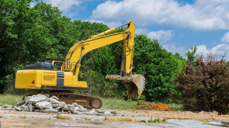 Koparka rozbija beton na placu, wokół zieleń i chmury na niebie.