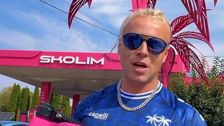 Mężczyzna w okularach przeciwsłonecznych i niebieskiej koszulce stoi przed stacją paliw Skolima na Mazurach.