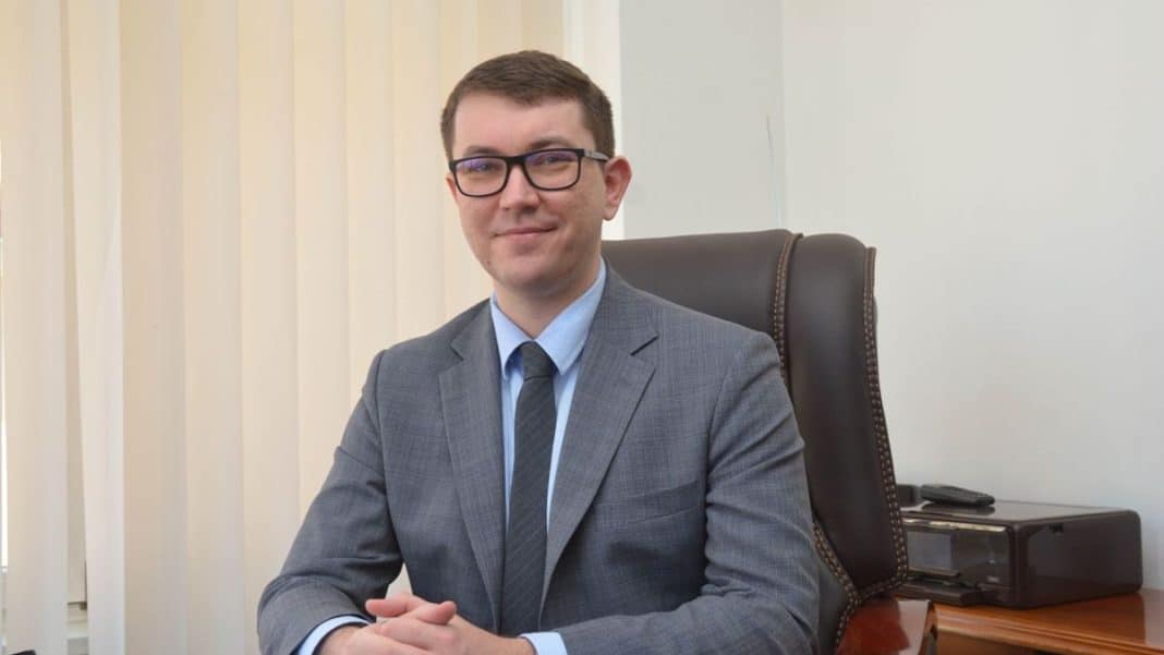 Mężczyzna w garniturze siedzi przy biurku w biurze, uśmiecha się do kamery.