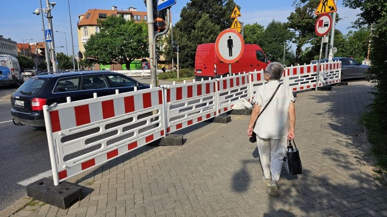 Pieszy przechodzi obok barierki i znaku zakazu wjazdu na ulicy w Olsztynie, wokół ruchliwa ulica z samochodami.
