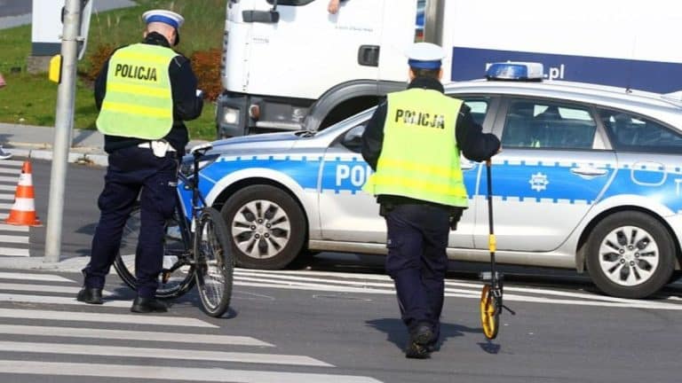 Policjanci na miejscu zdarzenia, przy samochodzie policyjnym i rowerze, na przejściu dla pieszych.