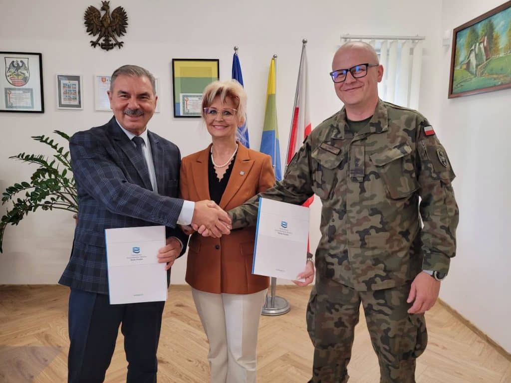 Trzech osób, w tym jeden w mundurze wojskowym, uśmiechają się i ściskają ręce podczas podpisywania dokumentów w pomieszczeniu z flagami i obrazami na ścianie.