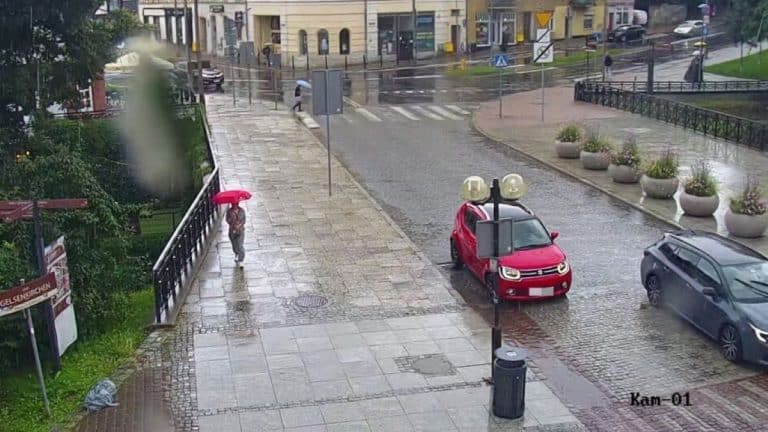 Widok na mokry, deszczowy chodnik i zaparkowany na moście czerwony samochód, obok pieszy z parasolem.