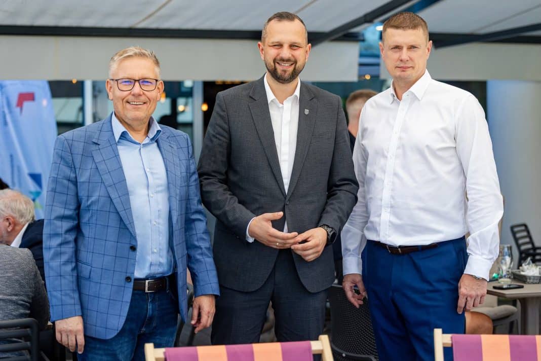 Trzech mężczyzn w formalnych ubraniach na spotkaniu, uśmiechają się do kamery, w tle inne osoby i elementy wnętrza.