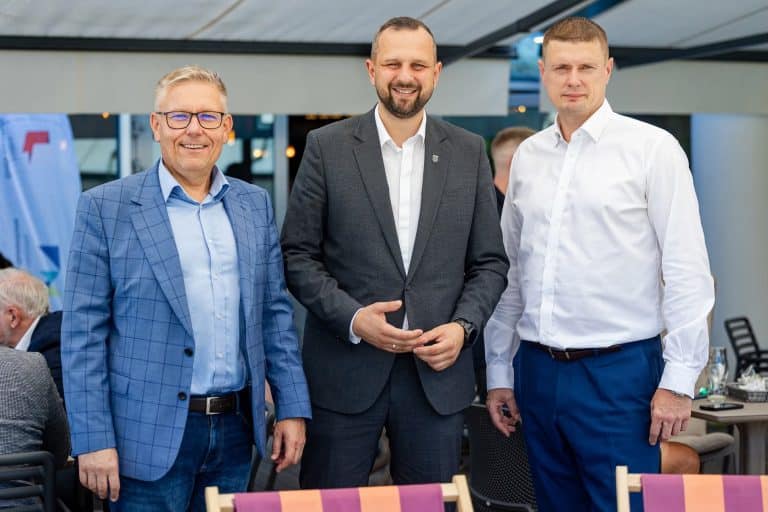 Trzech mężczyzn w formalnych ubraniach na spotkaniu, uśmiechają się do kamery, w tle inne osoby i elementy wnętrza.