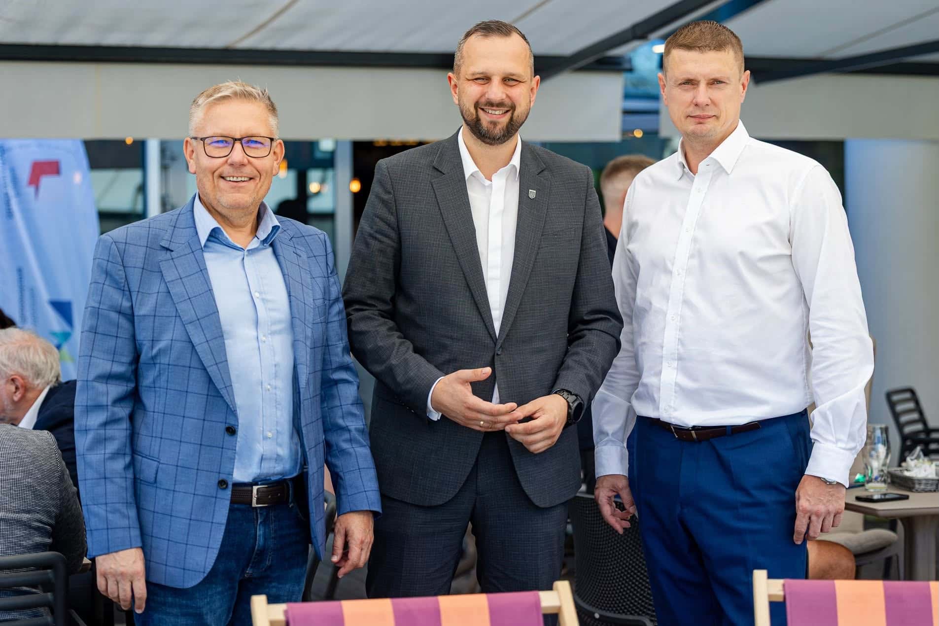 Trzech mężczyzn w formalnych ubraniach na spotkaniu, uśmiechają się do kamery, w tle inne osoby i elementy wnętrza.