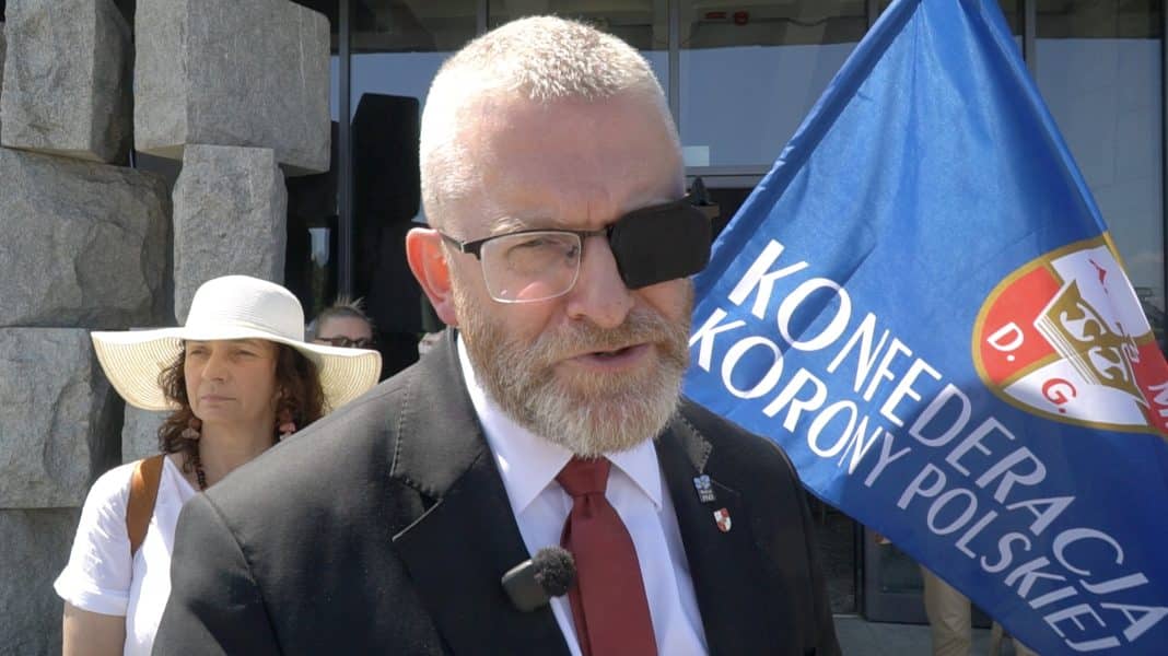 Mężczyzna z blizną na oku, trzymający flagę z napisem o konferencji, podczas wydarzenia na Polach Grunwaldzkich.