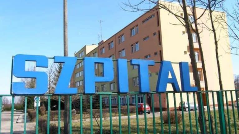 Zdjęcie budynku szpitala z dużym napisem 'SZPITAL' na ogrodzeniu przed wejściem.