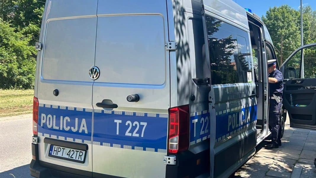 Policjant stoi przy oznakowanym pojeździe policyjnym na ulicy, w tle drzewa i niebo.