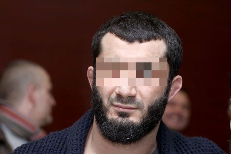 Mężczyzna z ciemną brodą i krótkimi włosami, z zasłoniętymi oczami, na tle innych osób w pomieszczeniu.