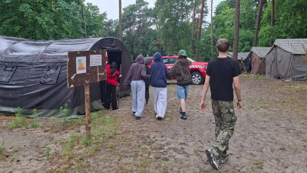 Grupa harcerzy w obozie, w tle namioty i budynek, podczas ewakuacji z powodu gwałtownej pogody.