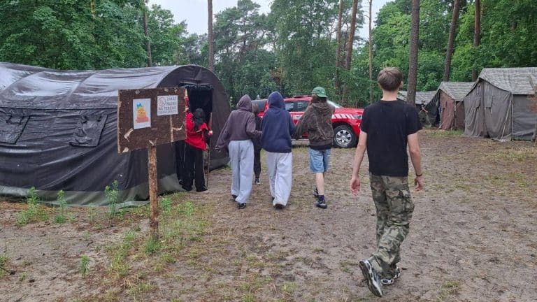 Grupa harcerzy w obozie, w tle namioty i budynek, podczas ewakuacji z powodu gwałtownej pogody.