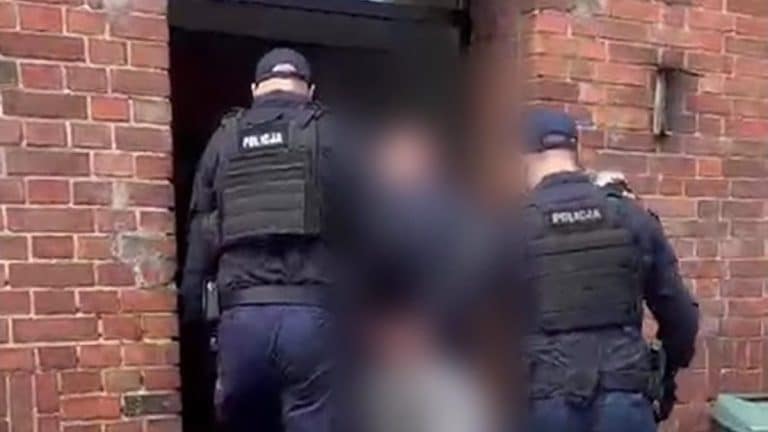 Policjanci zatrzymują podejrzanego mężczyznę w bloku, od tyłu, w czapkach i kamizelkach z napisem 'Policja'.