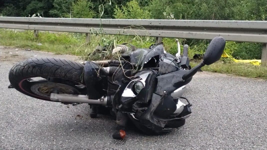 Zniszczony motocykl leży na drodze po wypadku, za nim barierka i zieleń.