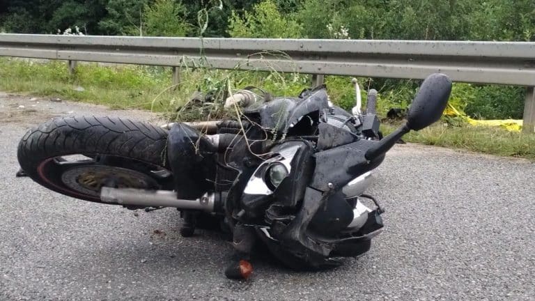 Zniszczony motocykl leży na drodze po wypadku, za nim barierka i zieleń.