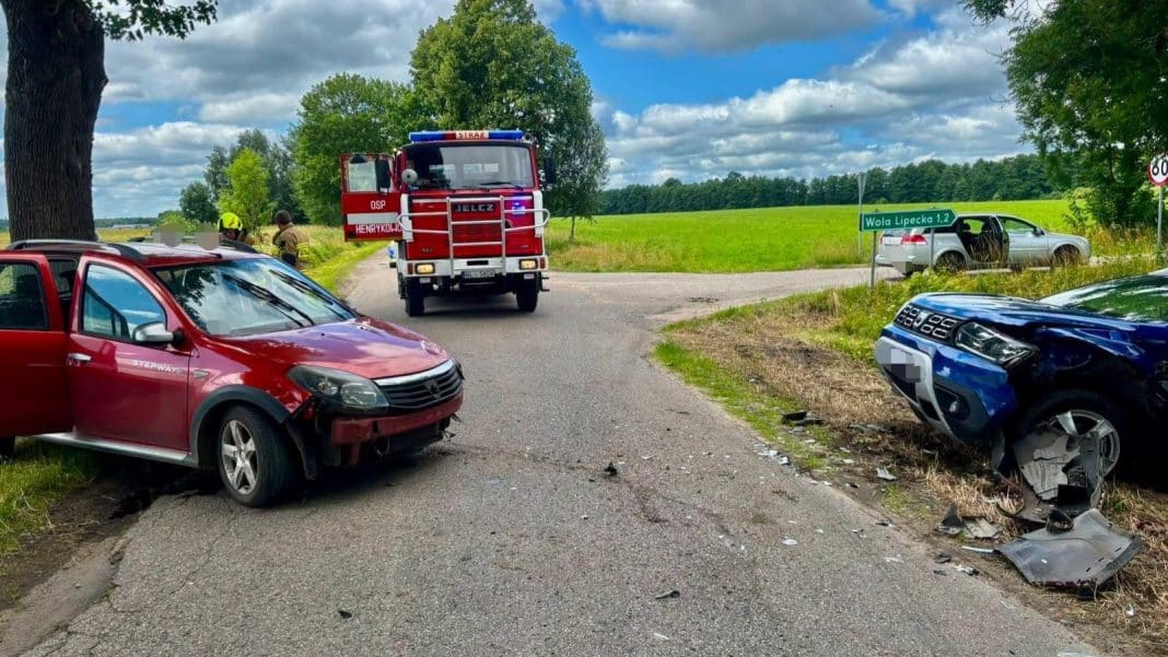 Zdjęcie przedstawia zderzenie dwóch samochodów marki Dacia na drodze, z pojazdami uszkodzonymi i służbami ratunkowymi na miejscu.