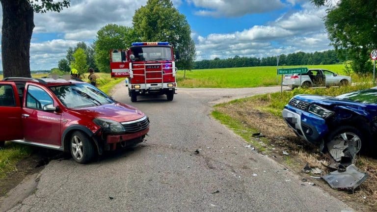 Zdjęcie przedstawia zderzenie dwóch samochodów marki Dacia na drodze, z pojazdami uszkodzonymi i służbami ratunkowymi na miejscu.