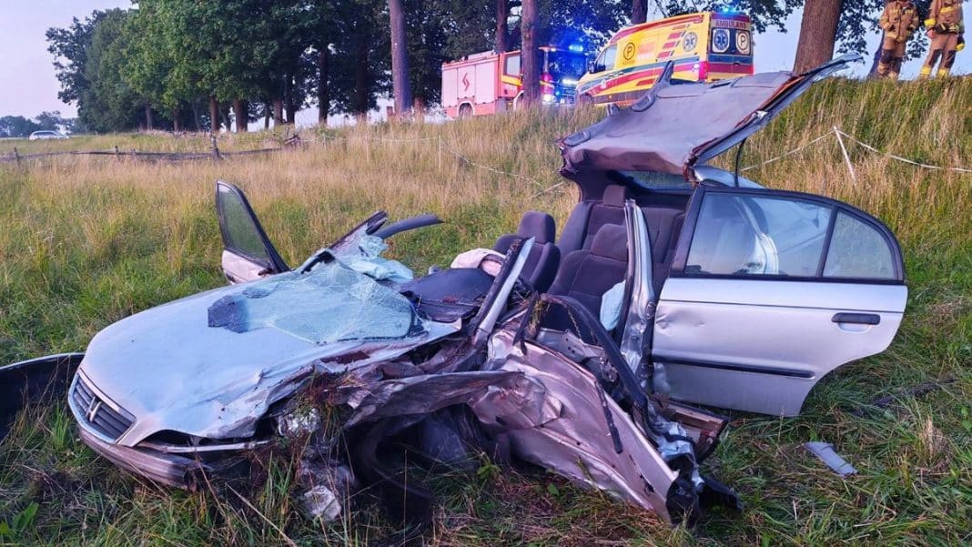 Rozbity samochód osobowy po wypadku z widocznymi uszkodzeniami, na tle służb ratunkowych i straży pożarnej.