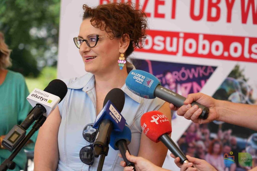 Kobieta w okularach i kolorowych kolczykach udziela wywiadu podczas konferencji, otoczona mikrofonami mediów.