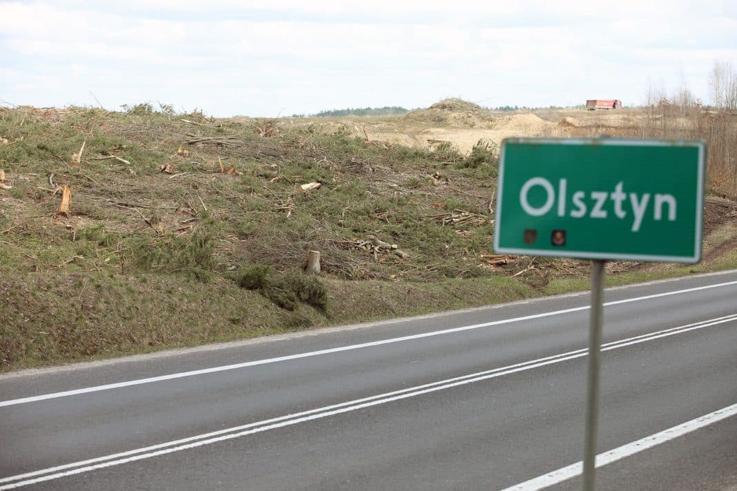 Zdjęcie drogi z tablicą Olsztyn i wycinkiem lasu lub terenu z wyciętymi drzewami.