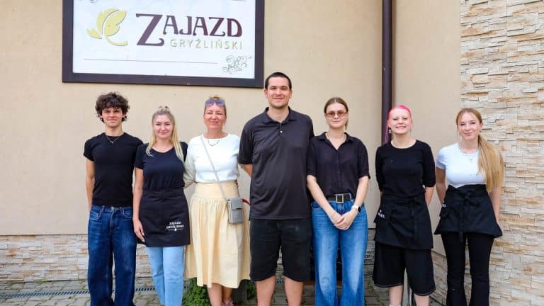 Grupa ludzi stojąca przed budynkiem z tablicą Zajazd Gryźliński, wśród nich cztery kobiety i mężczyzna.