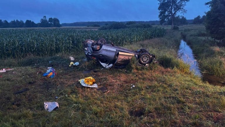 Rozbity samochód na boku na poboczu drogi, wokół śmieci i rozrzucone przedmioty, w tle pola i drzewa.