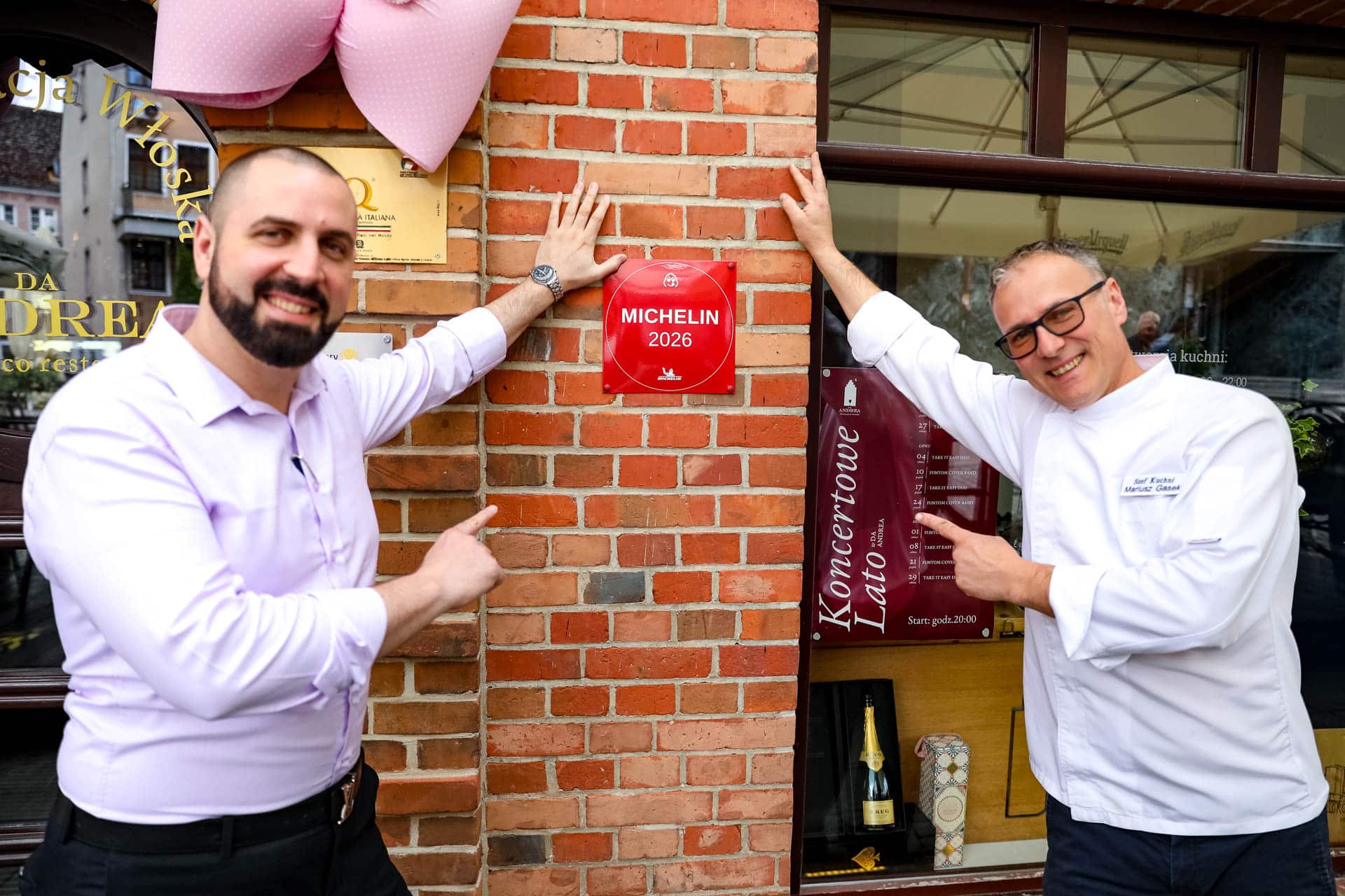 Dwóch mężczyzn stojących przed restauracją, wskazujących na certyfikat Michelin Bib Gourmand na ścianie.