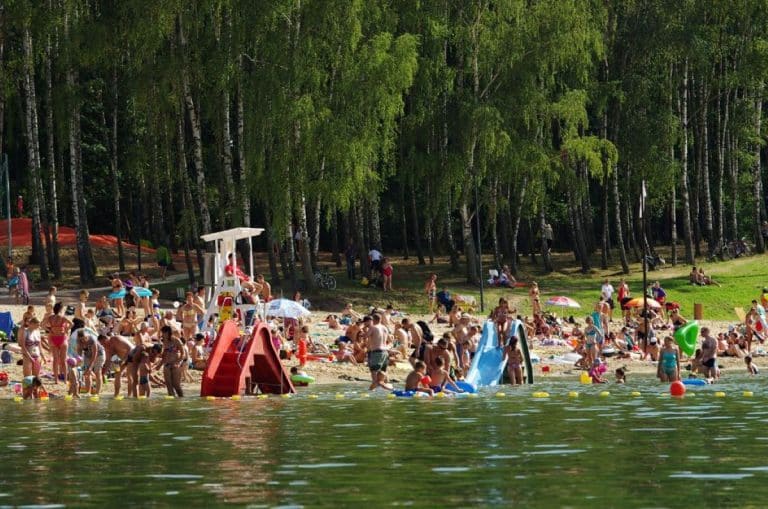 Ludzie na plaży nad jeziorem, kąpią się i odpoczywają w słoneczny dzień, wokół zielone drzewa.