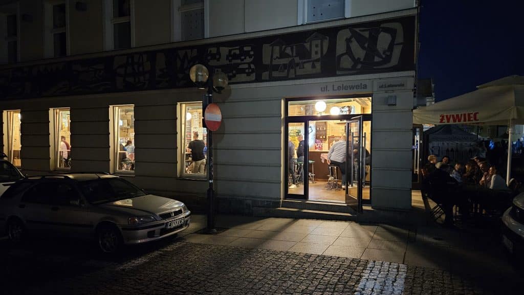 Widok na lokal gastronomiczny na starówce w Olsztynie wieczorem, z widocznymi oknami i grupami ludzi na zewnątrz.