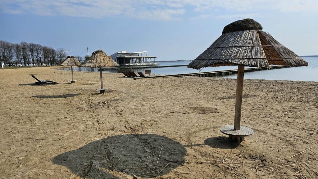 Pusty, piaszczysty plaża z parasolami z trzciny i leżakami, widok na jezioro i nowoczesny budynek w tle.