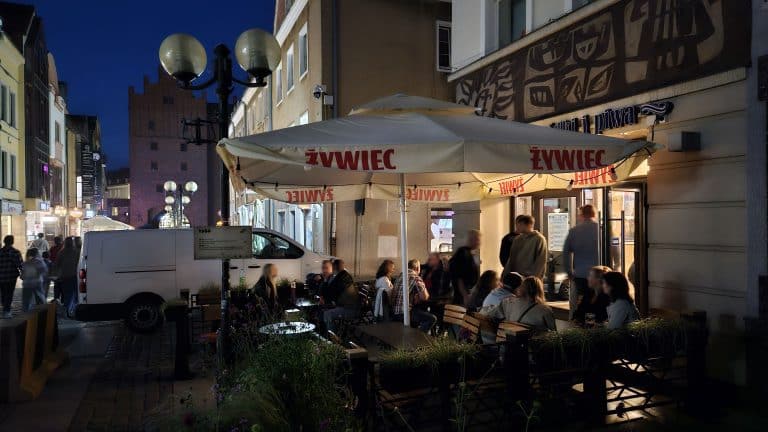 Wieczorne zdjęcie lokalu gastronomicznego na starówce z ludźmi na zewnątrz pod parasolami.