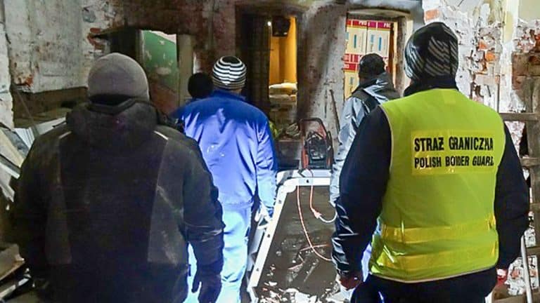 Grupa funkcjonariuszy Straży Granicznej w pomieszczeniu podczas kontroli, w tle widoczne ściany z cegieł i sprzęt.