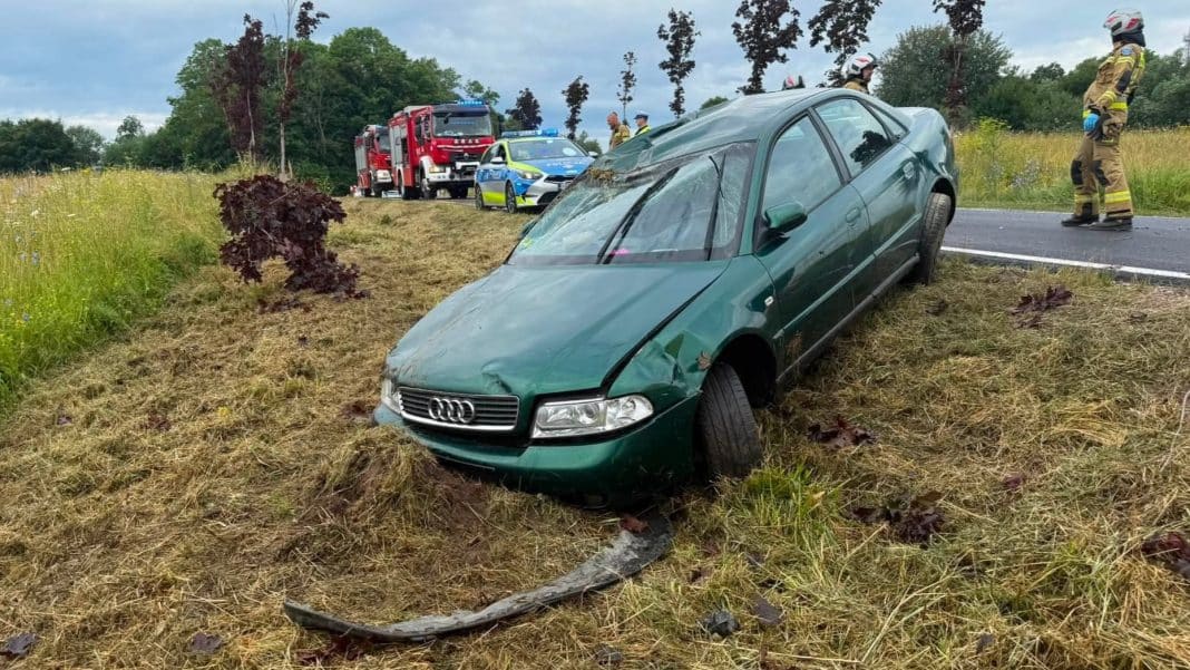 Samochód marki Audi po wypadku leży na poboczu, w tle służby ratunkowe i straż pożarna.