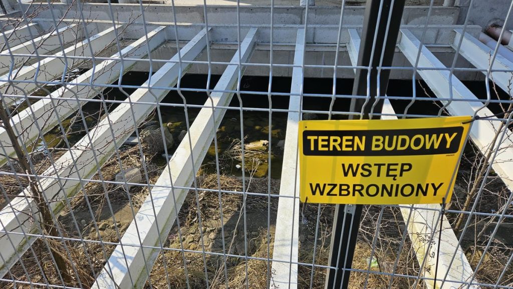 Zabezpieczony teren budowy z tablicą zakazu wjazdu, widoczna konstrukcja i woda w wykopie.