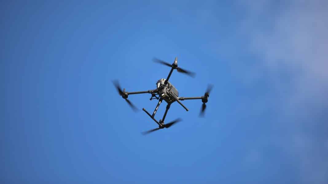 Dron latający nad niebem, widoczny z dołu, na tle błękitnego nieba.