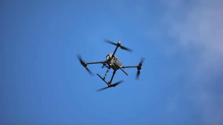 Dron latający nad niebem, widoczny z dołu, na tle błękitnego nieba.