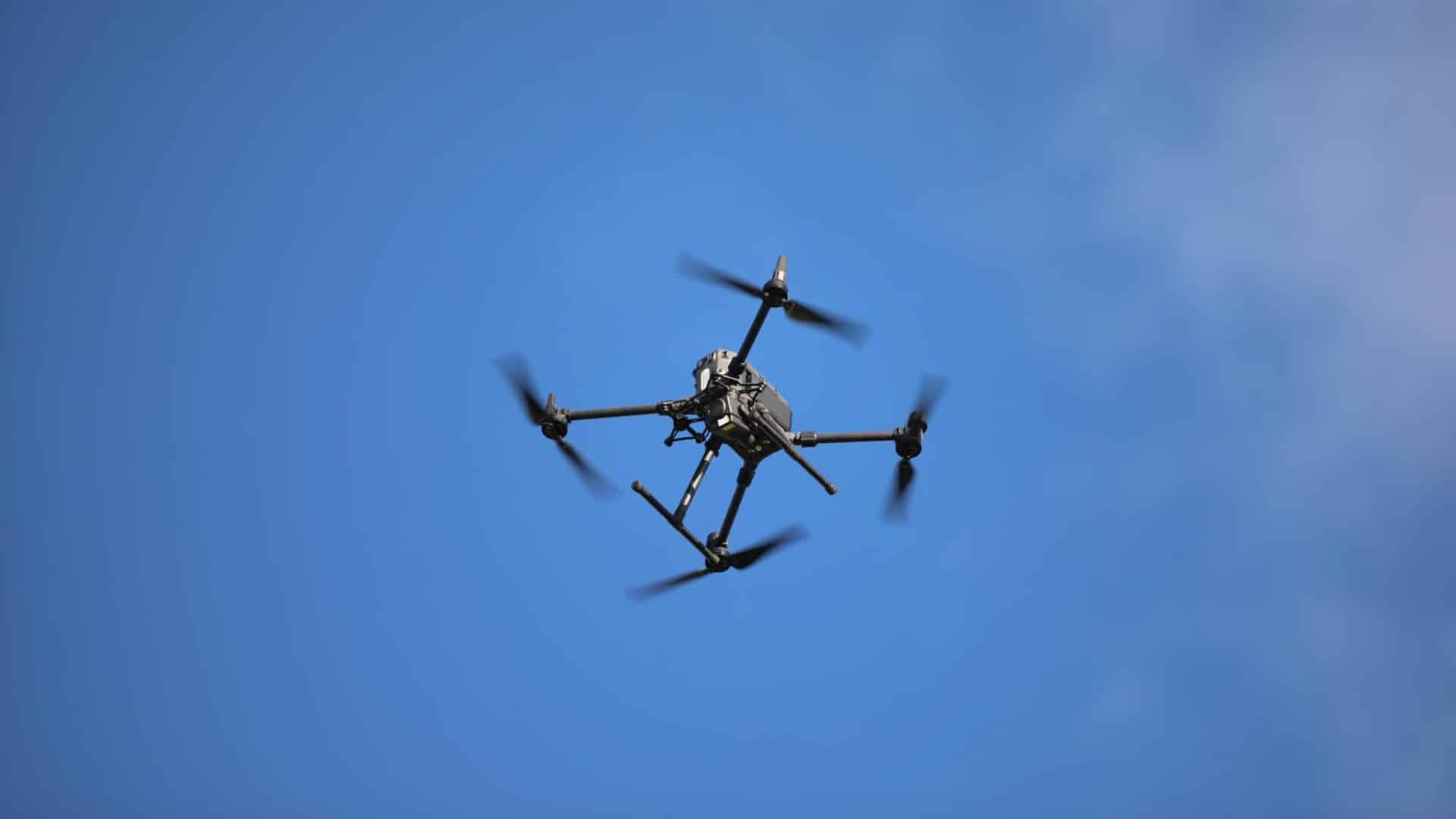 Dron latający nad niebem, widoczny z dołu, na tle błękitnego nieba.