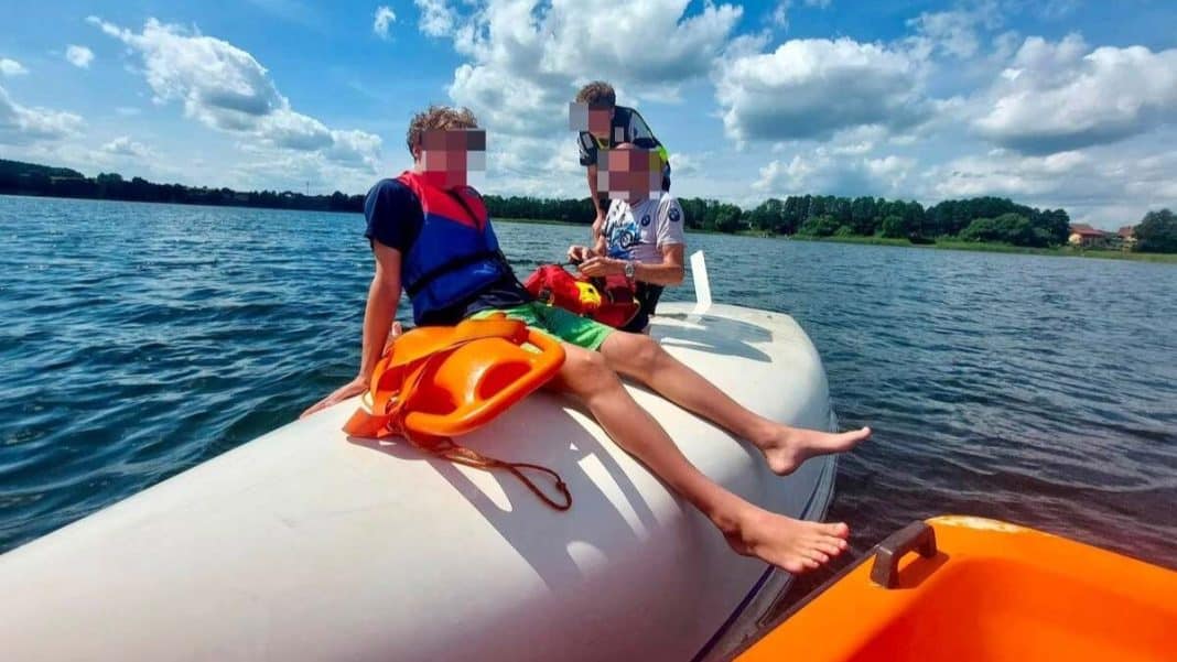 Dzieci na pontonie na jeziorze, jedna z nich z kamizelką ratunkową, w tle woda i chmury na niebie.