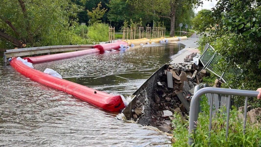 Woda zalała teren wokół parku, uszkodzona bariera zbiornika retencyjnego, widoczne uszkodzenia i rozlewisko.