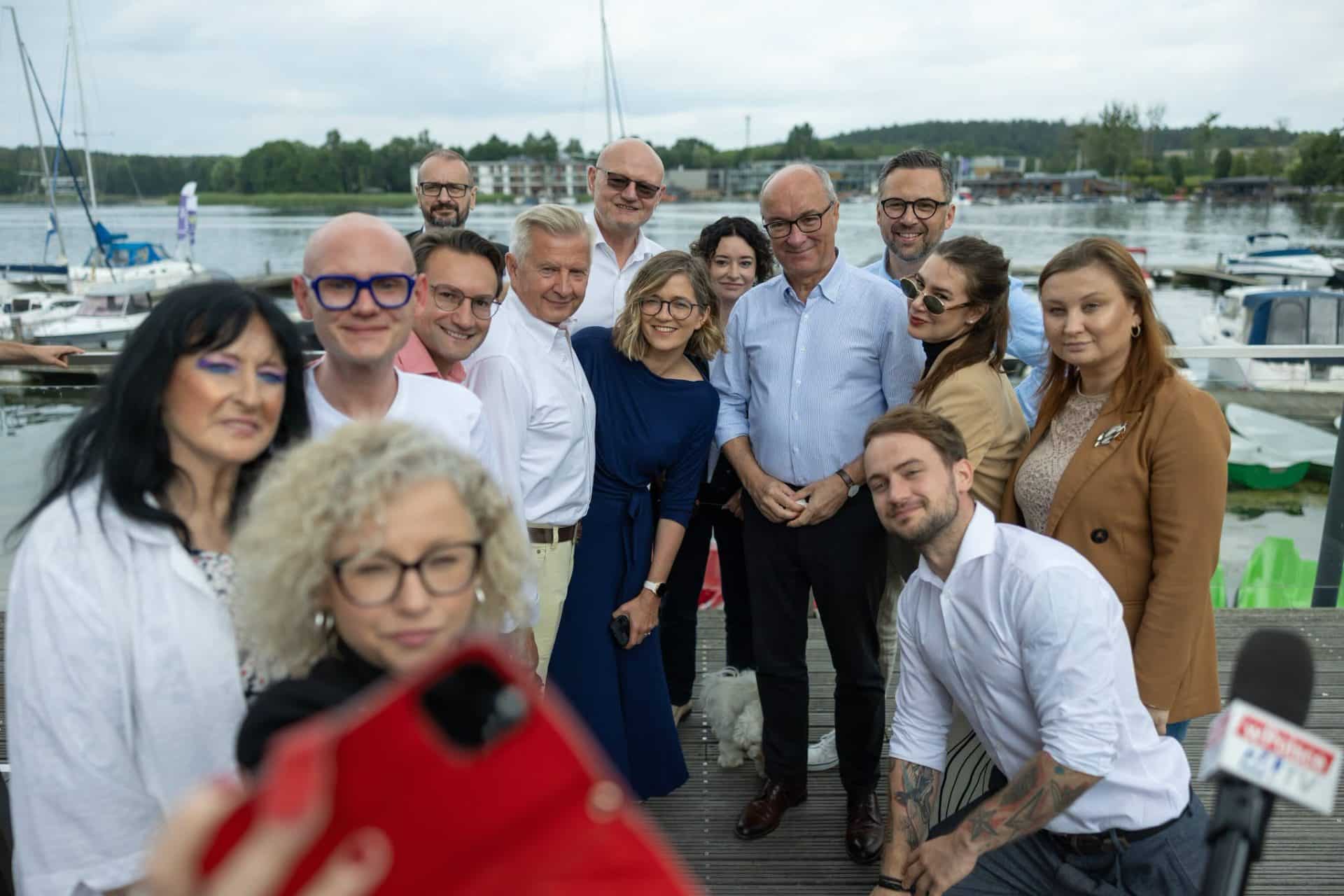 Grupa polityków i mieszkańców nad jeziorem Ukiel w Olsztynie, w tle łodzie i budynki nad wodą.