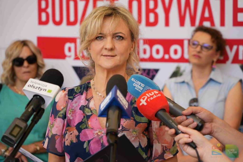 Kobieta w kwiatowej bluzce podczas konferencji z mikrofonami w tle.