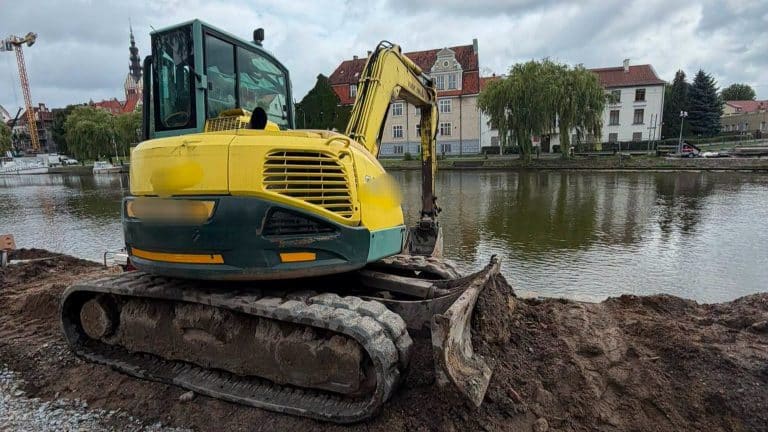 Żółta koparka na placu budowy przy rzece, w tle budynki i drzewa.