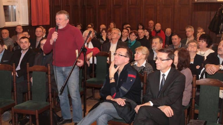 Mężczyzna z mikrofonem przemawia do zgromadzonej publiczności w sali konferencyjnej, za nim siedzi grupa słuchaczy.